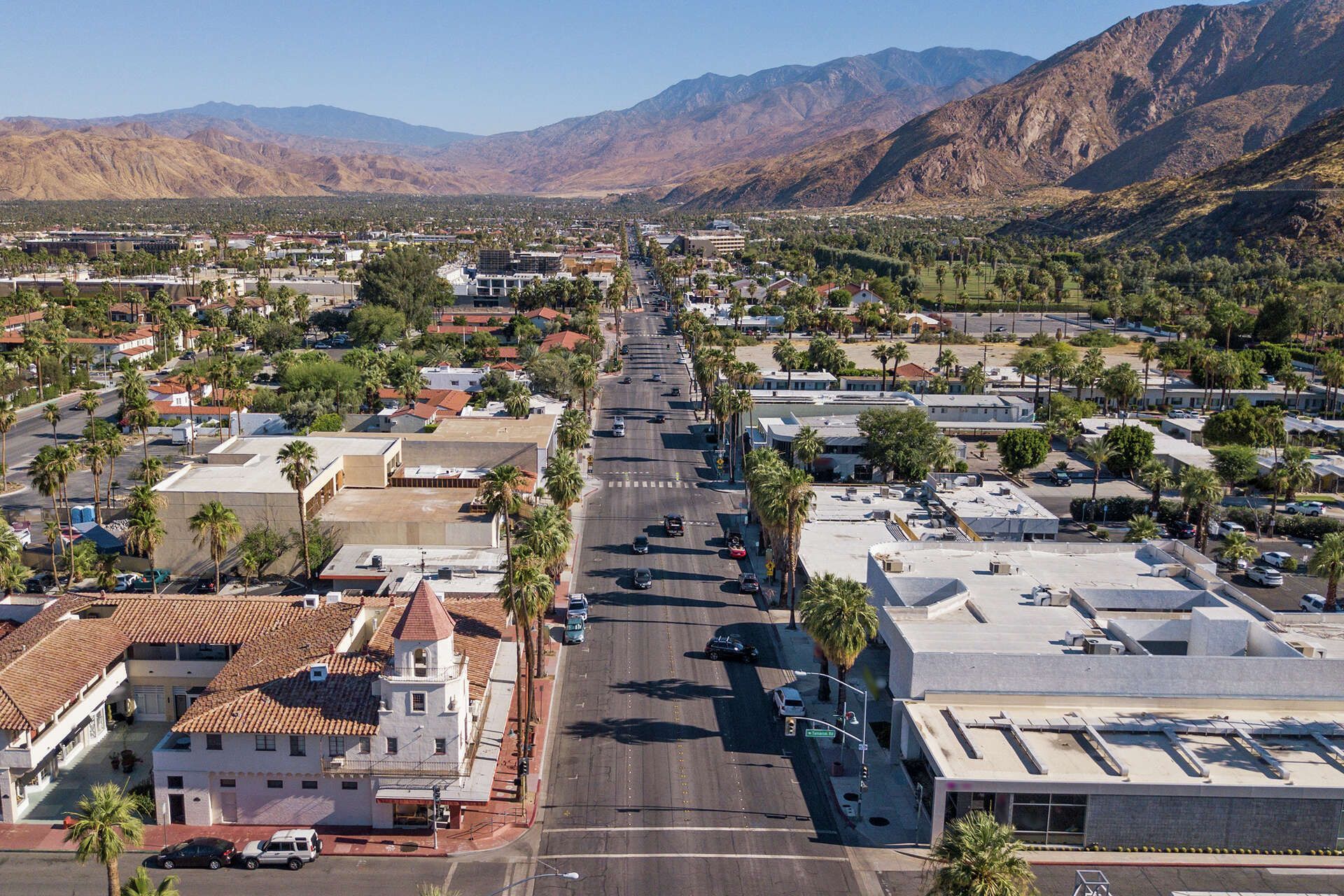 Aerial view of a street in Palm Springs, California, with buildings, palm trees, and mountains in the background.