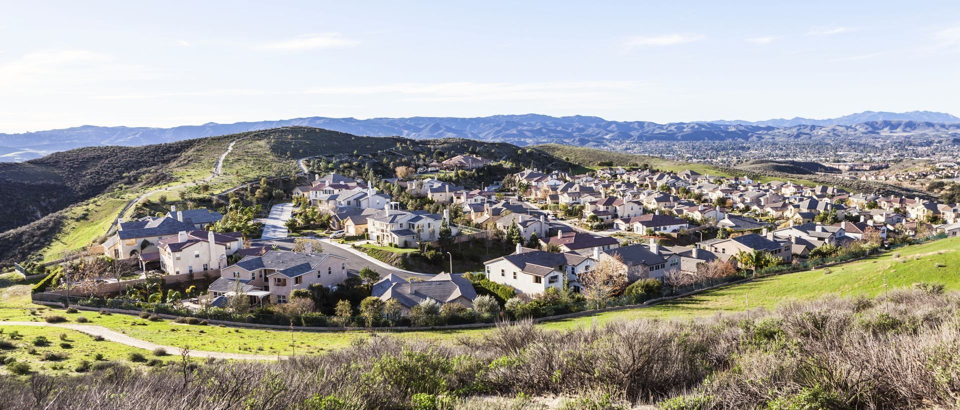 A sprawling neighborhood of houses nestled on a green hillside, with rolling hills and a clear blue sky in the background.