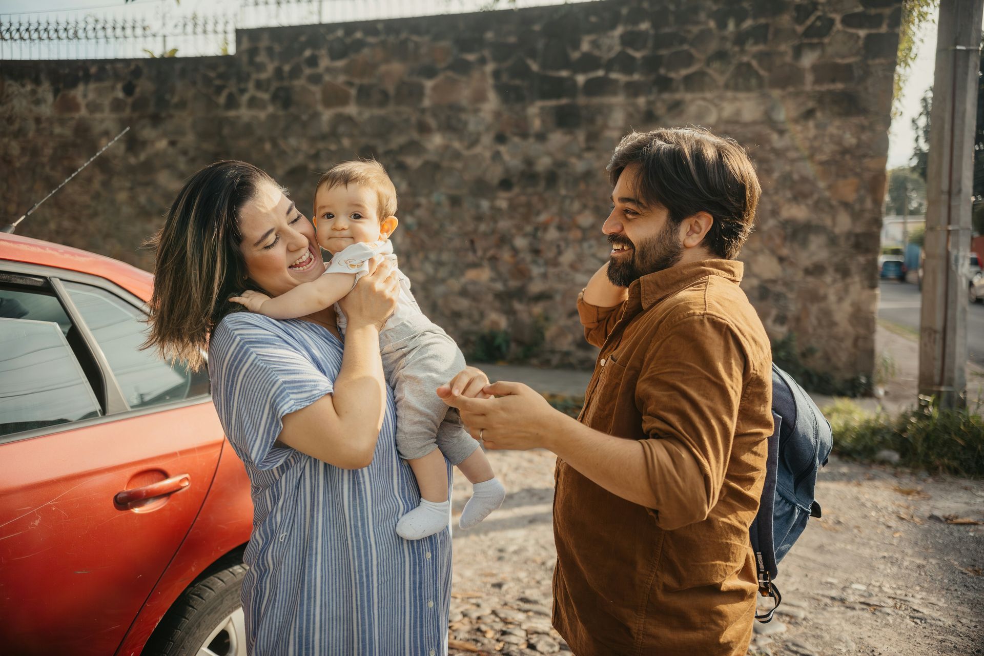 A man and a woman are holding a baby in front of a red car.
