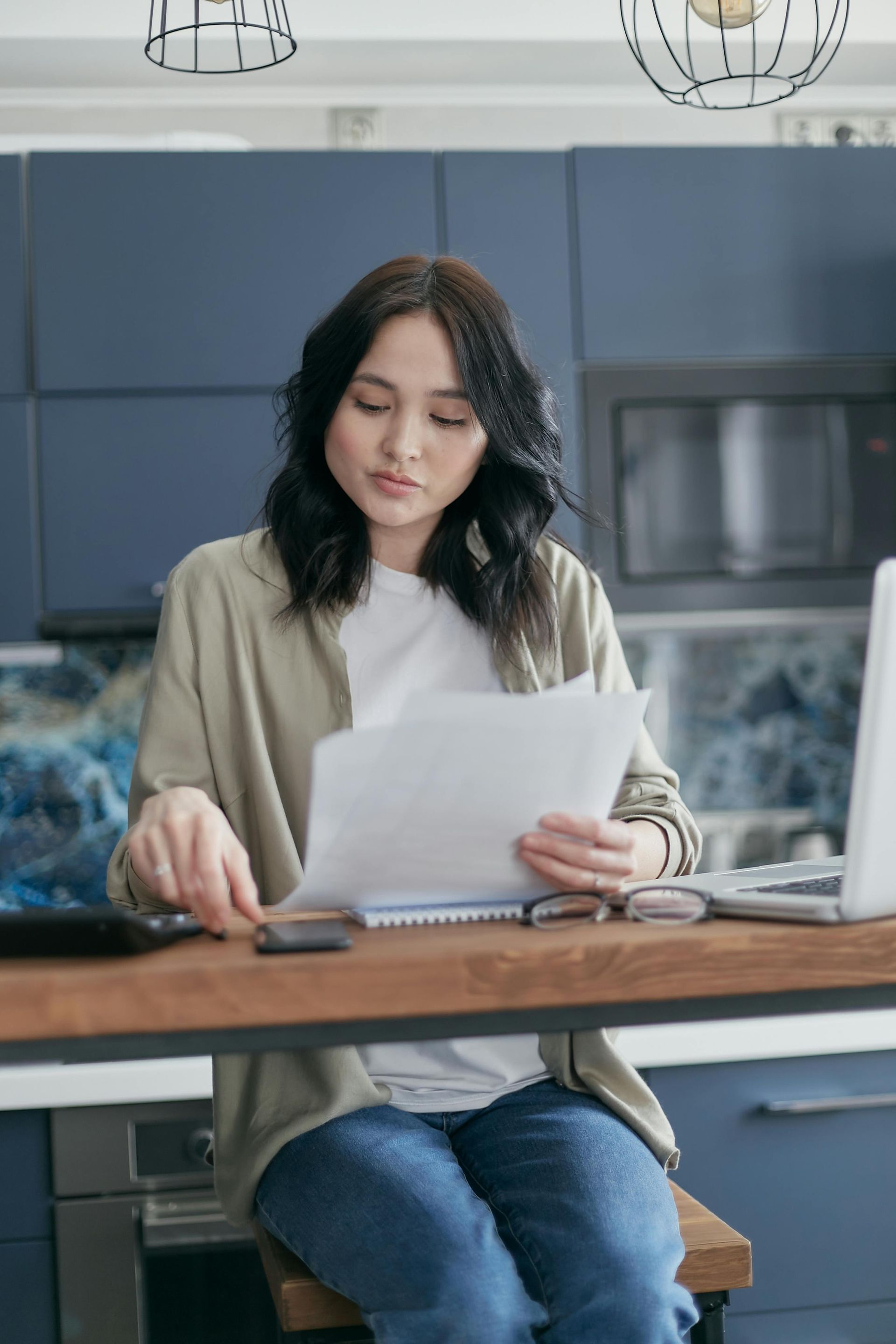 Woman in kitchen reviews papers, calculator, phone, and laptop on counter.