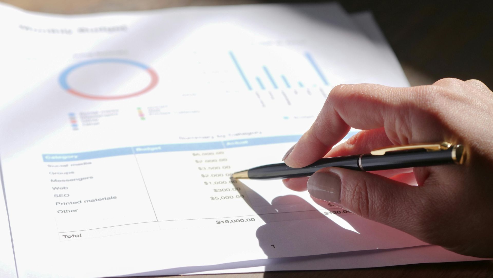 Hand holding pen points at financial data on paper.