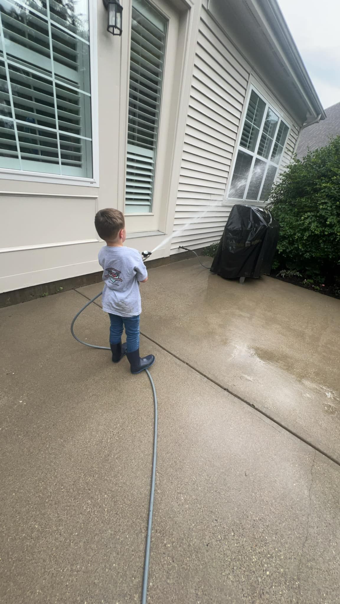 A little boy is standing on a sidewalk holding a hose in front of a house.