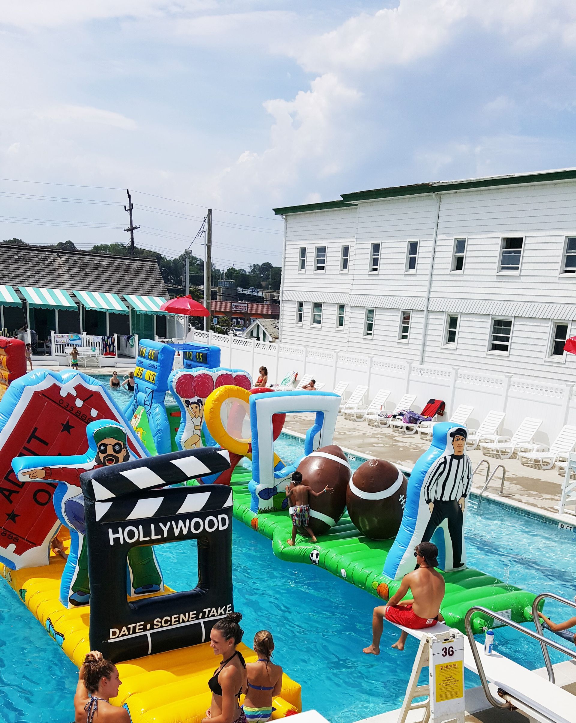 An inflatable obstacle course sits in a bright blue swimming pool, featuring footballs and a Hollywood-themed sign.