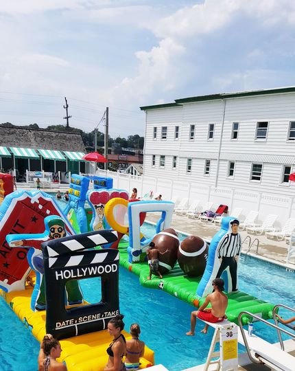 An inflatable obstacle course sits in a bright blue swimming pool, featuring footballs and a Hollywood-themed sign.