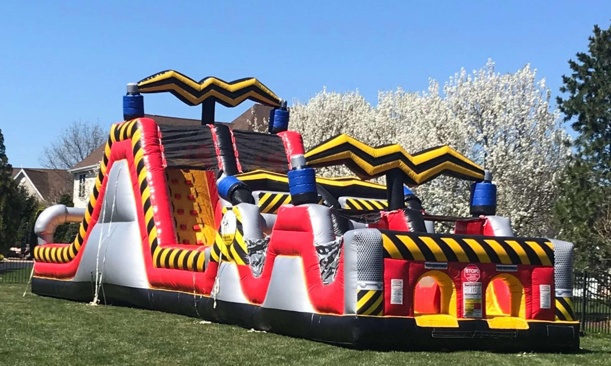 Red, black, and yellow inflatable obstacle course with tunnels and a climbing wall set up on a grassy lawn.