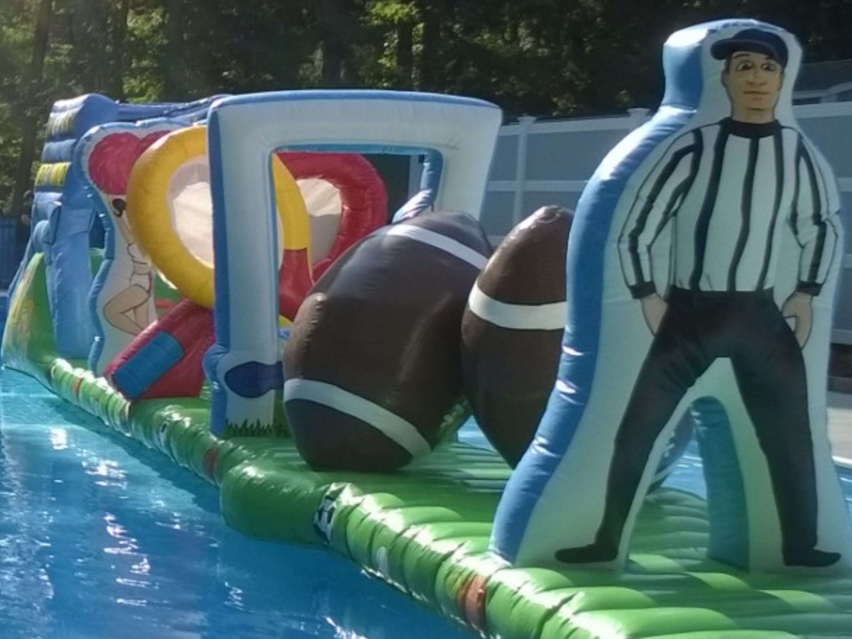 An inflatable football-themed obstacle course sits on a pool, featuring a referee cutout and football shapes.