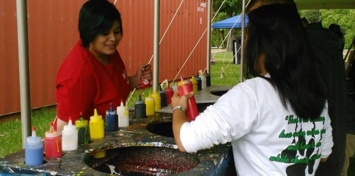 Two people work at an outdoor snow cone stand, using various colorful syrup bottles to prepare icy treats.