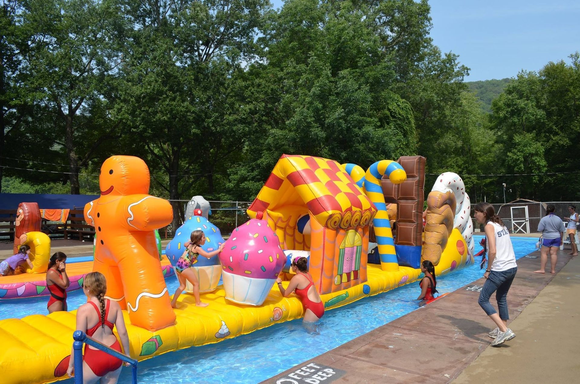 Children play on a colorful, candy-themed inflatable obstacle course floating in a pool outdoors.