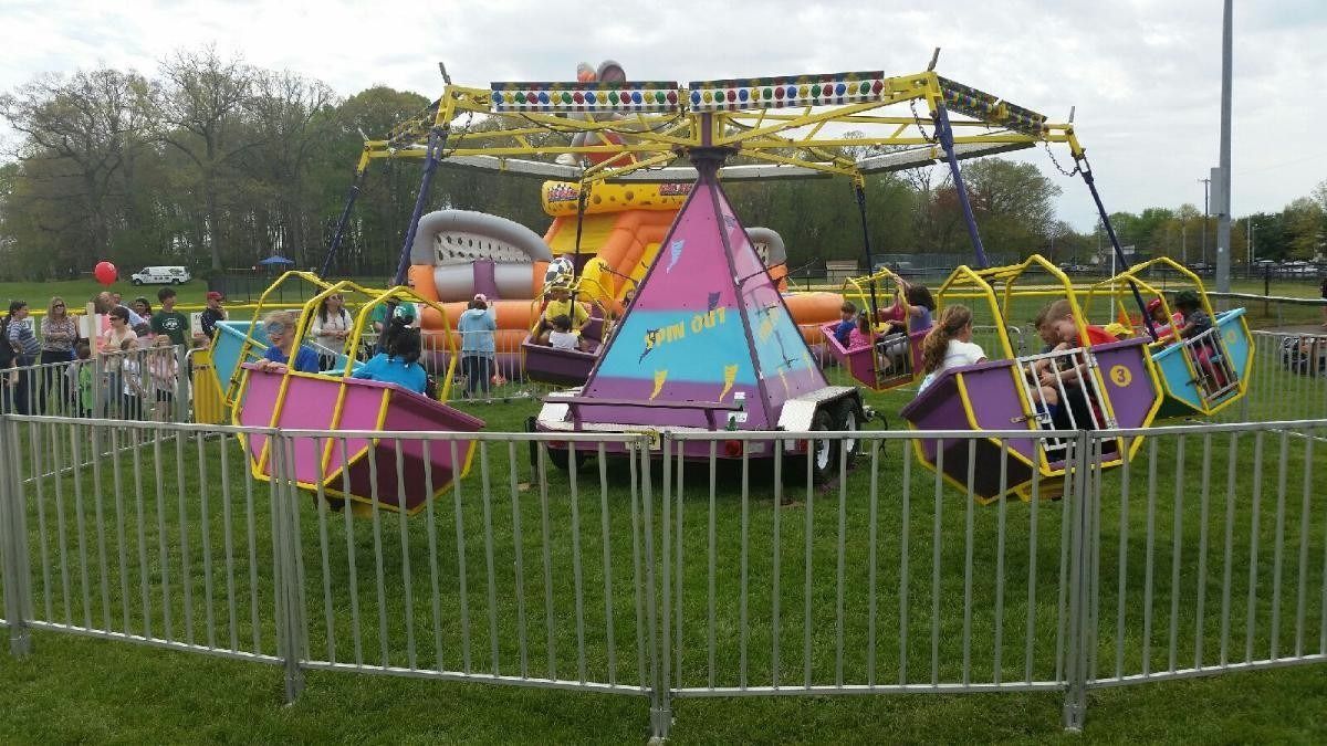 A colorful spinning swing ride for children operates on a grassy field, surrounded by a metal crowd-control fence.