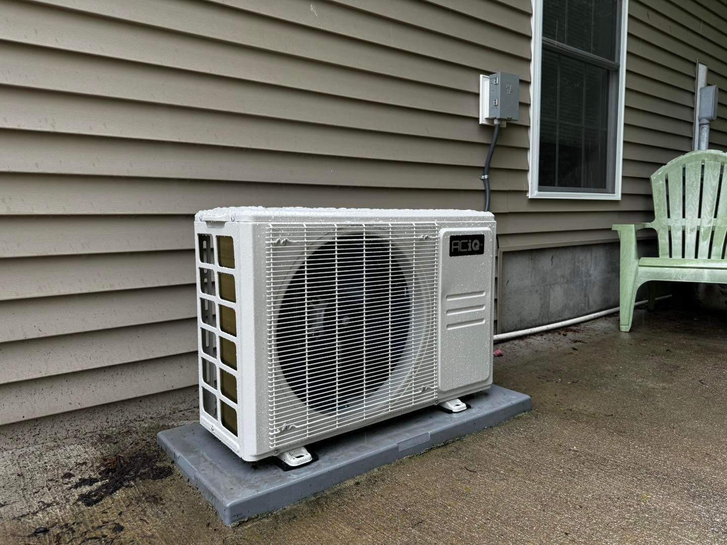 A white mini-split air conditioning unit sits on a grey concrete pad against the tan siding of a house.