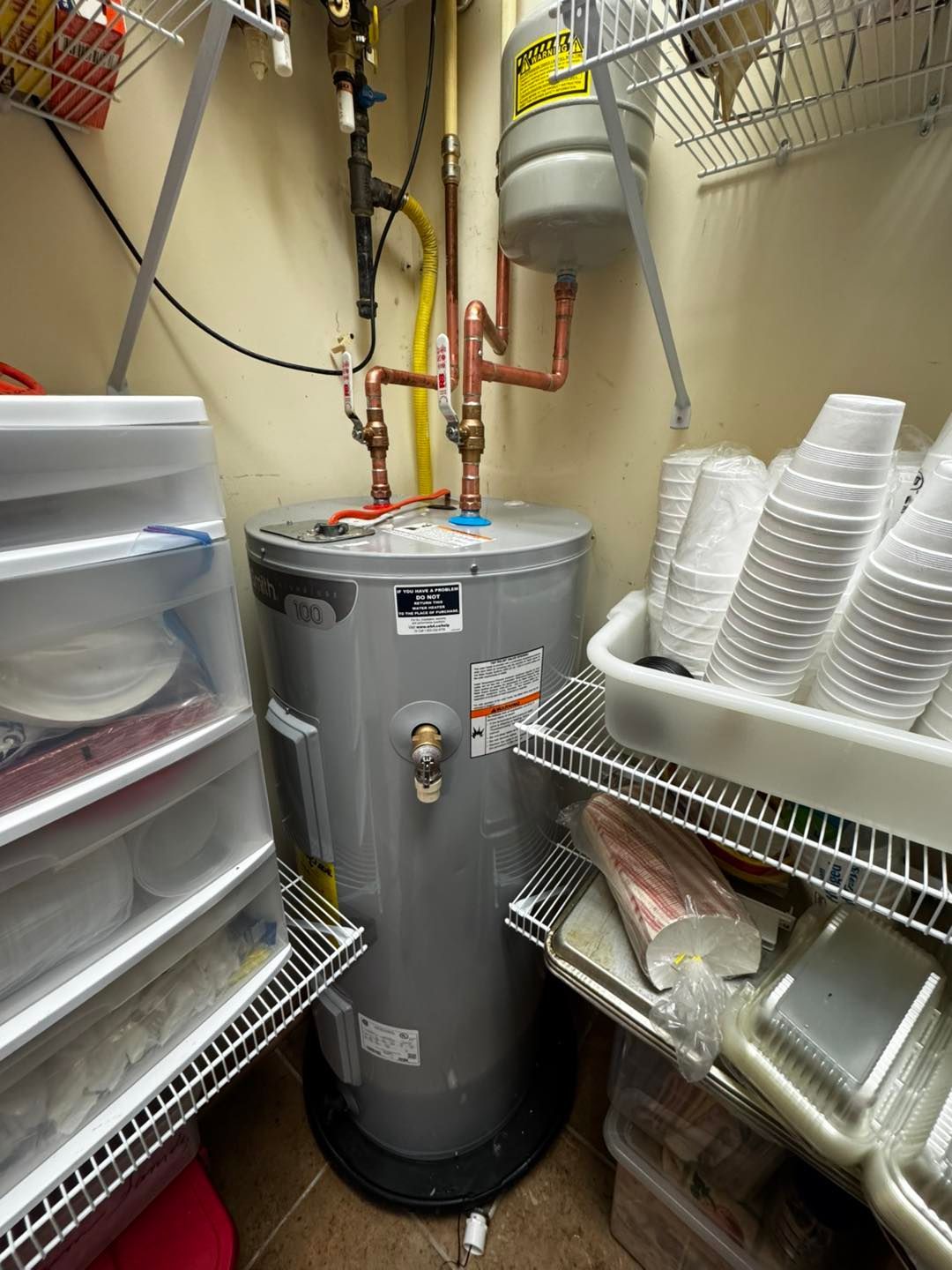 A gray electric water heater installed in a utility closet with storage shelves and stacked disposable cups.