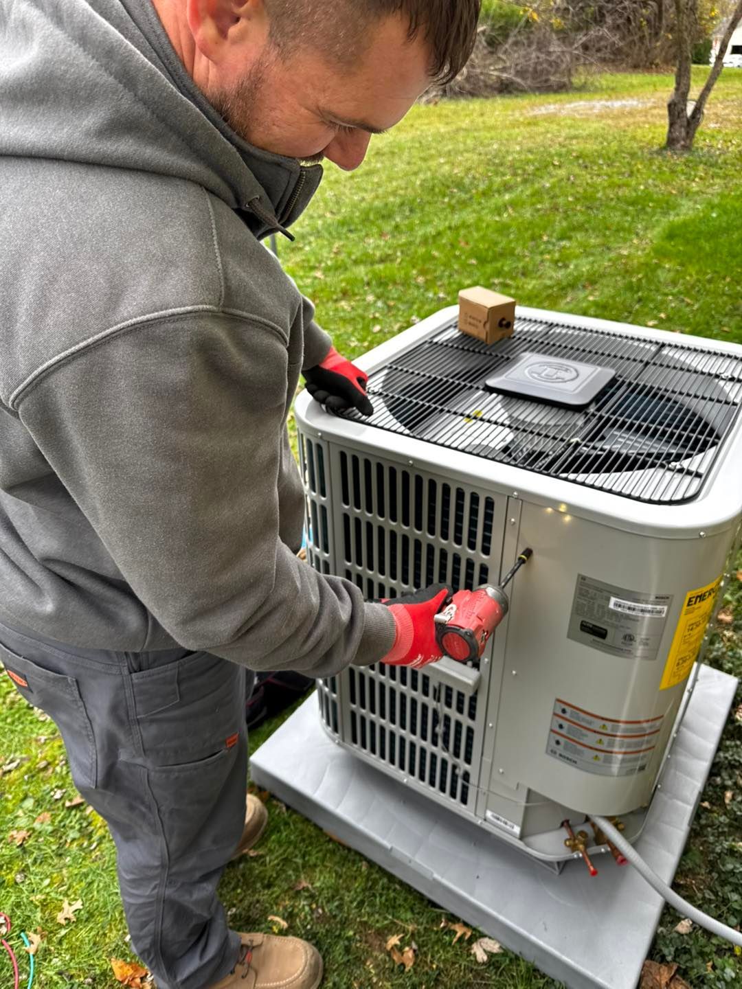 A technician in a gray hoodie uses a power drill to open an outdoor residential air conditioning unit on a grassy lawn.