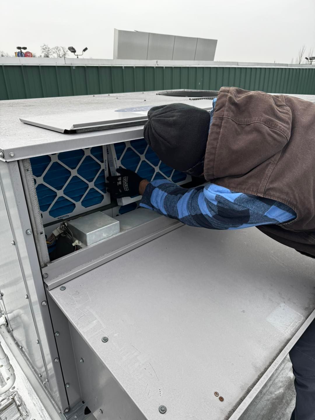 A worker in a brown vest and blue flannel shirt replaces a blue air filter inside an outdoor rooftop HVAC unit.