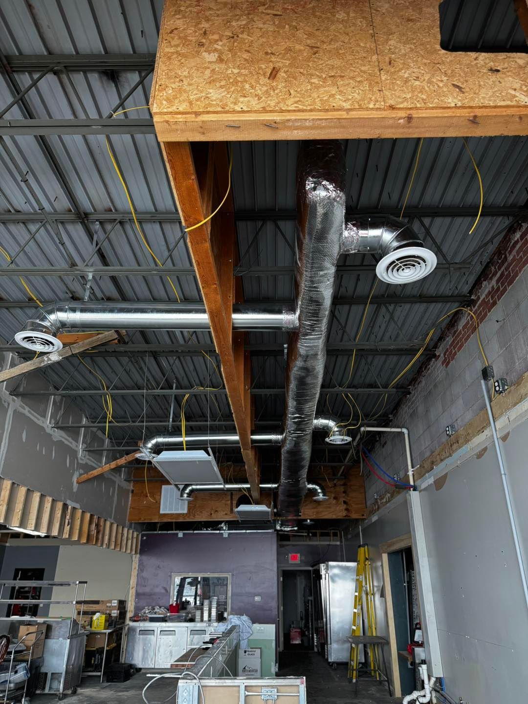 An interior view of a room under construction, featuring exposed ceiling rafters, ductwork, and hanging vents.