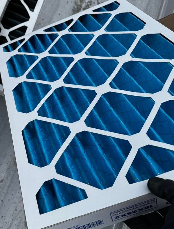 A person in a black glove holds a new, blue pleated air filter with a white diamond-pattern frame.
