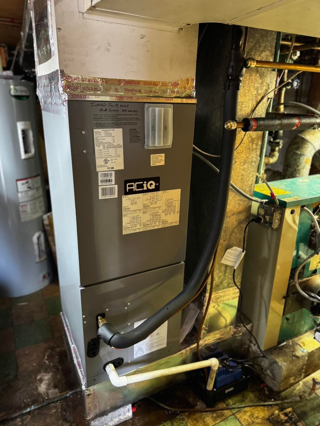 A gray HVAC air handler unit standing in a basement, connected to a drain line and condensate pump.