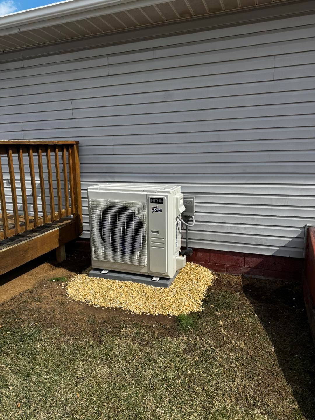 A beige outdoor heat pump unit sits on a gravel pad against the gray vinyl siding of a house next to a wooden deck.