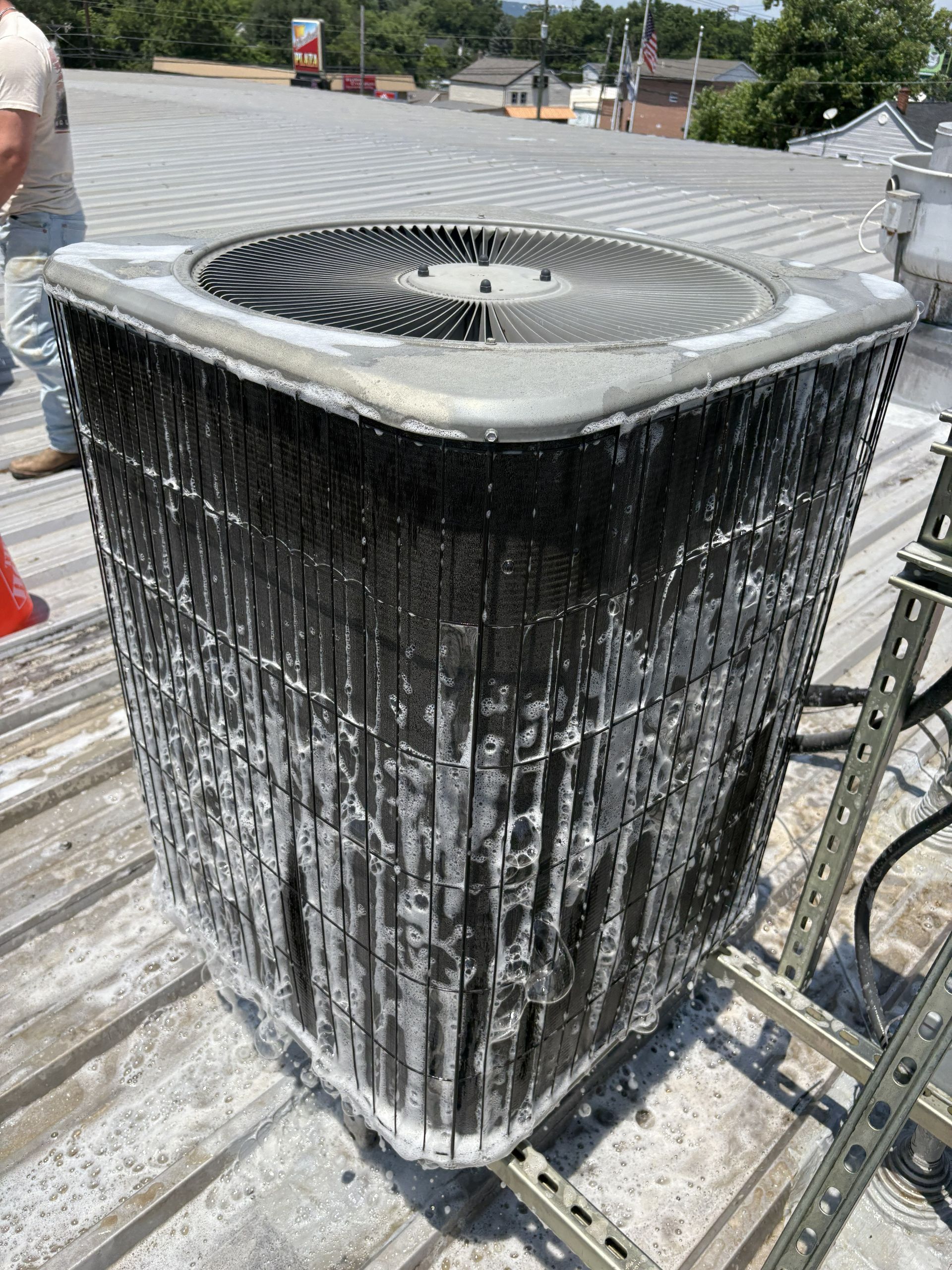 A technician works on an outdoor air conditioning unit with a heavily clogged and dirty condenser coil on a metal roof.