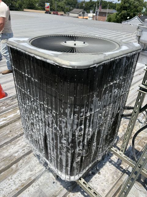 Two outdoor HVAC units mounted on a concrete pad against a white, corrugated metal wall under a small window.