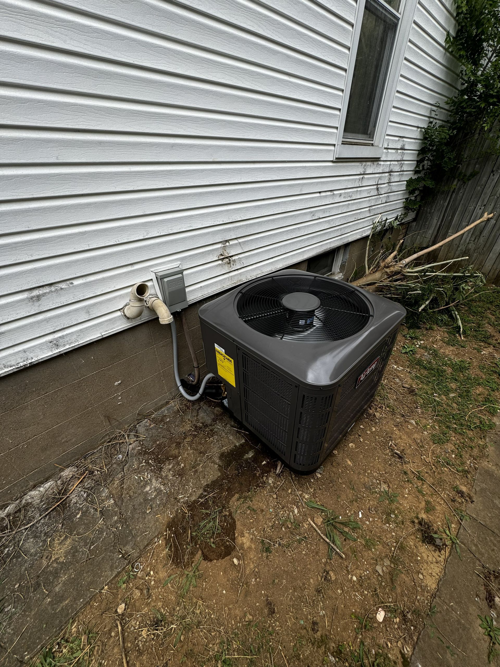 A dark gray outdoor air conditioning unit sits on the ground next to a white house with vinyl siding.