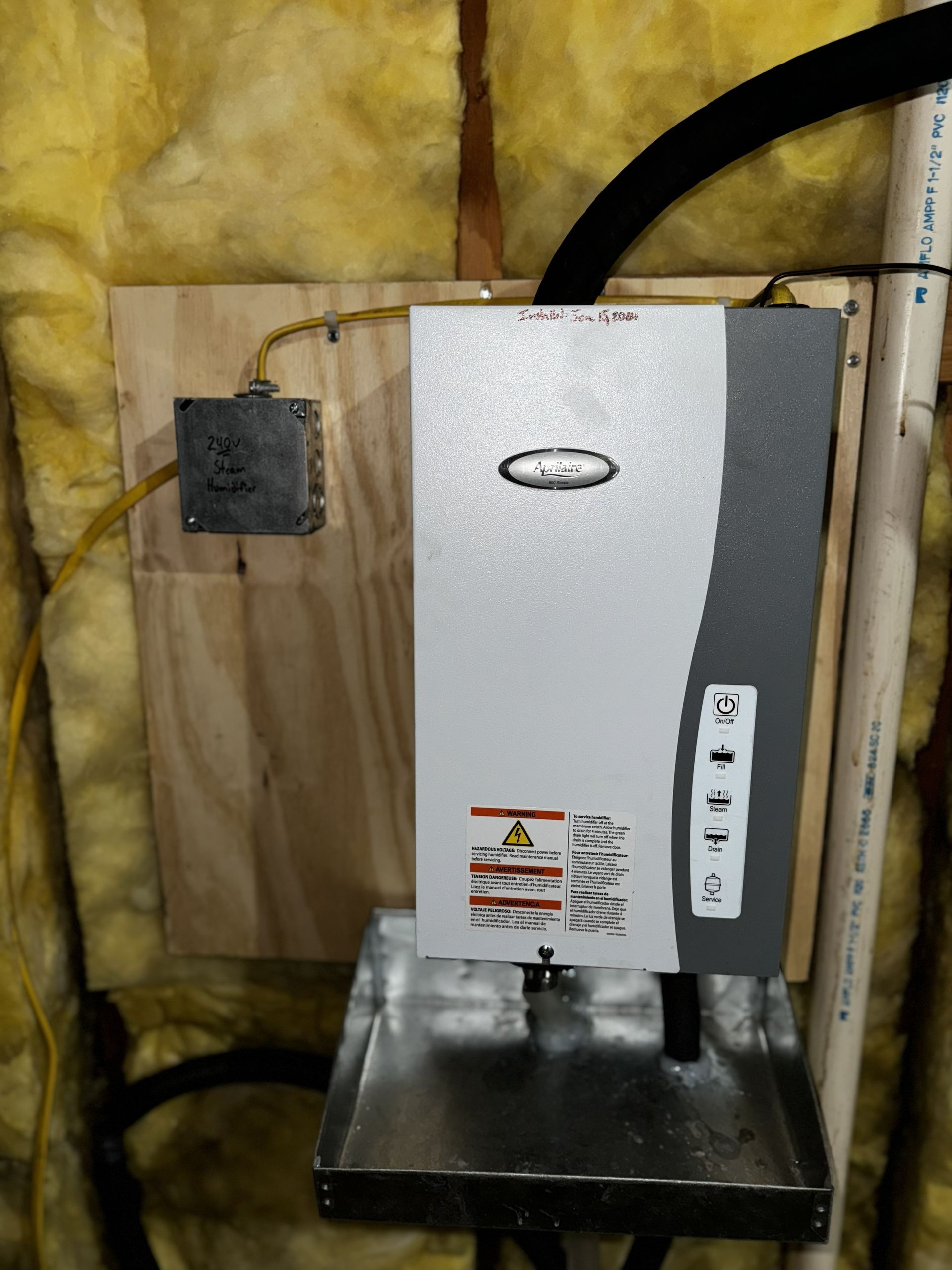 An electric tankless water heater mounted on a wooden board against insulated walls, with a drip pan underneath.