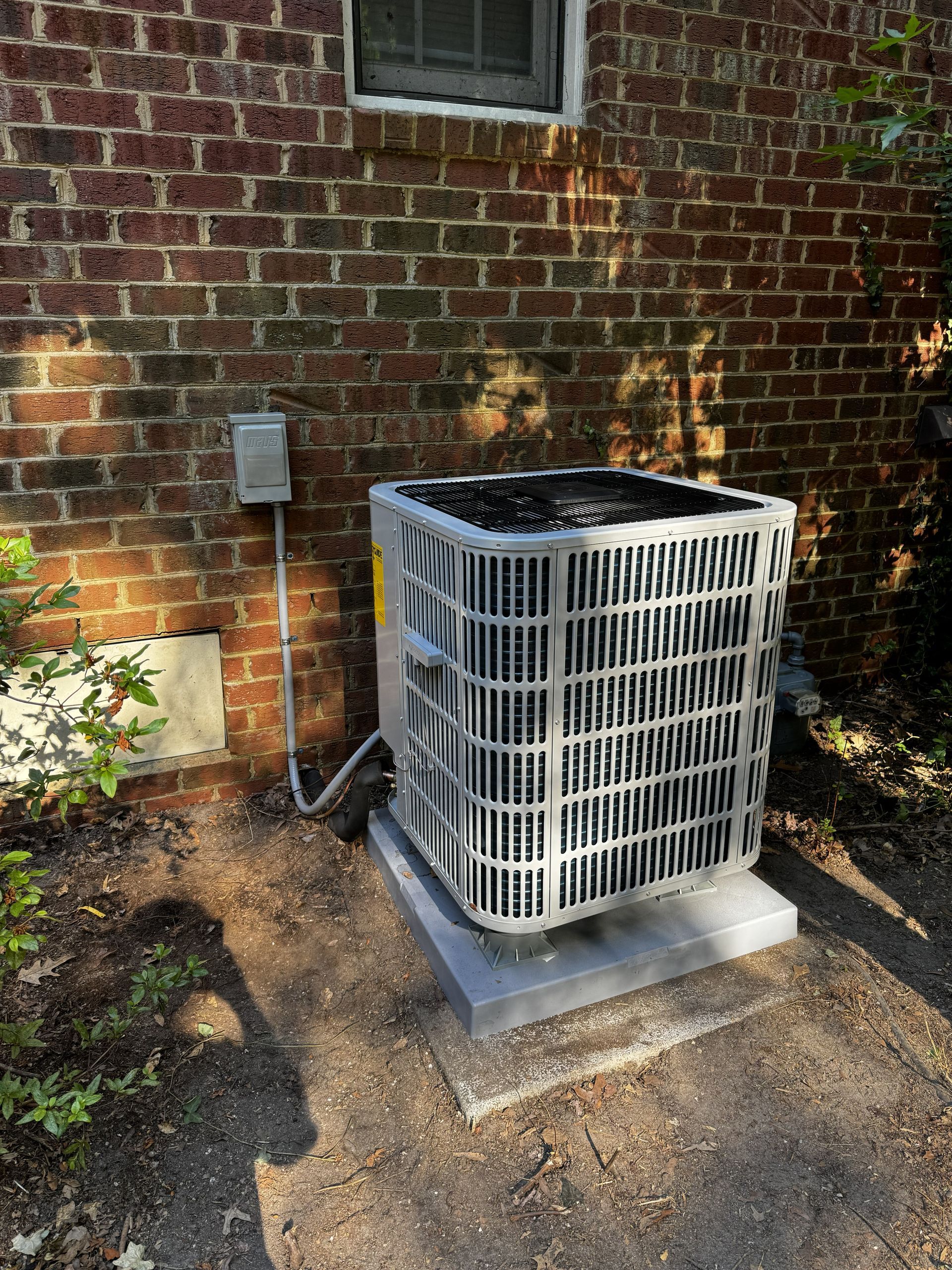 An outdoor central air conditioning unit sits on a concrete pad against a brick wall beneath a small window.