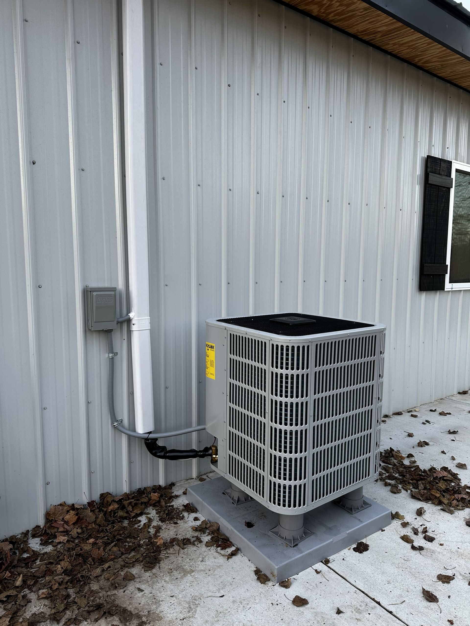 An outdoor HVAC condenser unit sits on a concrete pad next to a light gray metal wall with a white downspout.