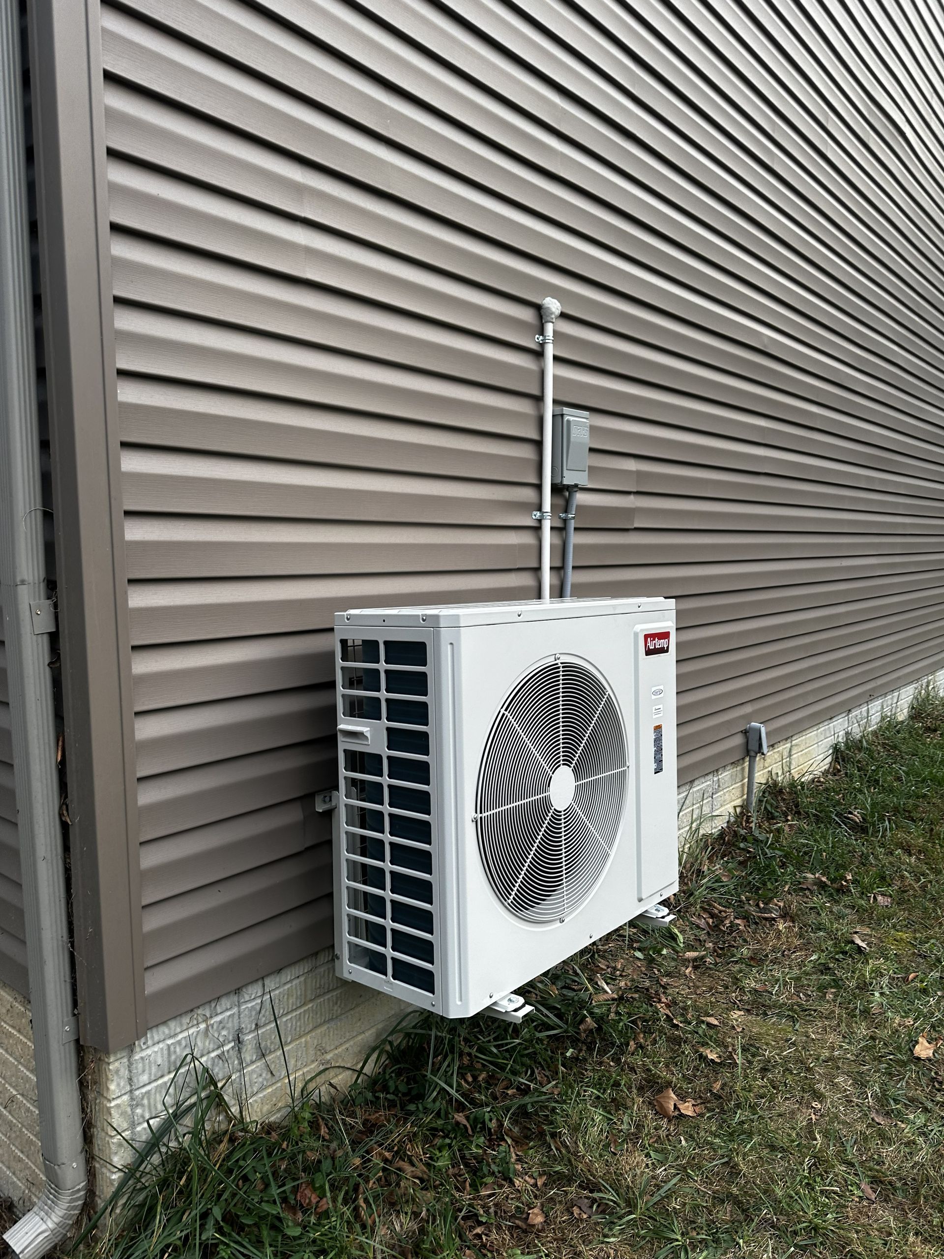 A white outdoor mini-split heat pump unit mounted on the brown vinyl siding of a house exterior.