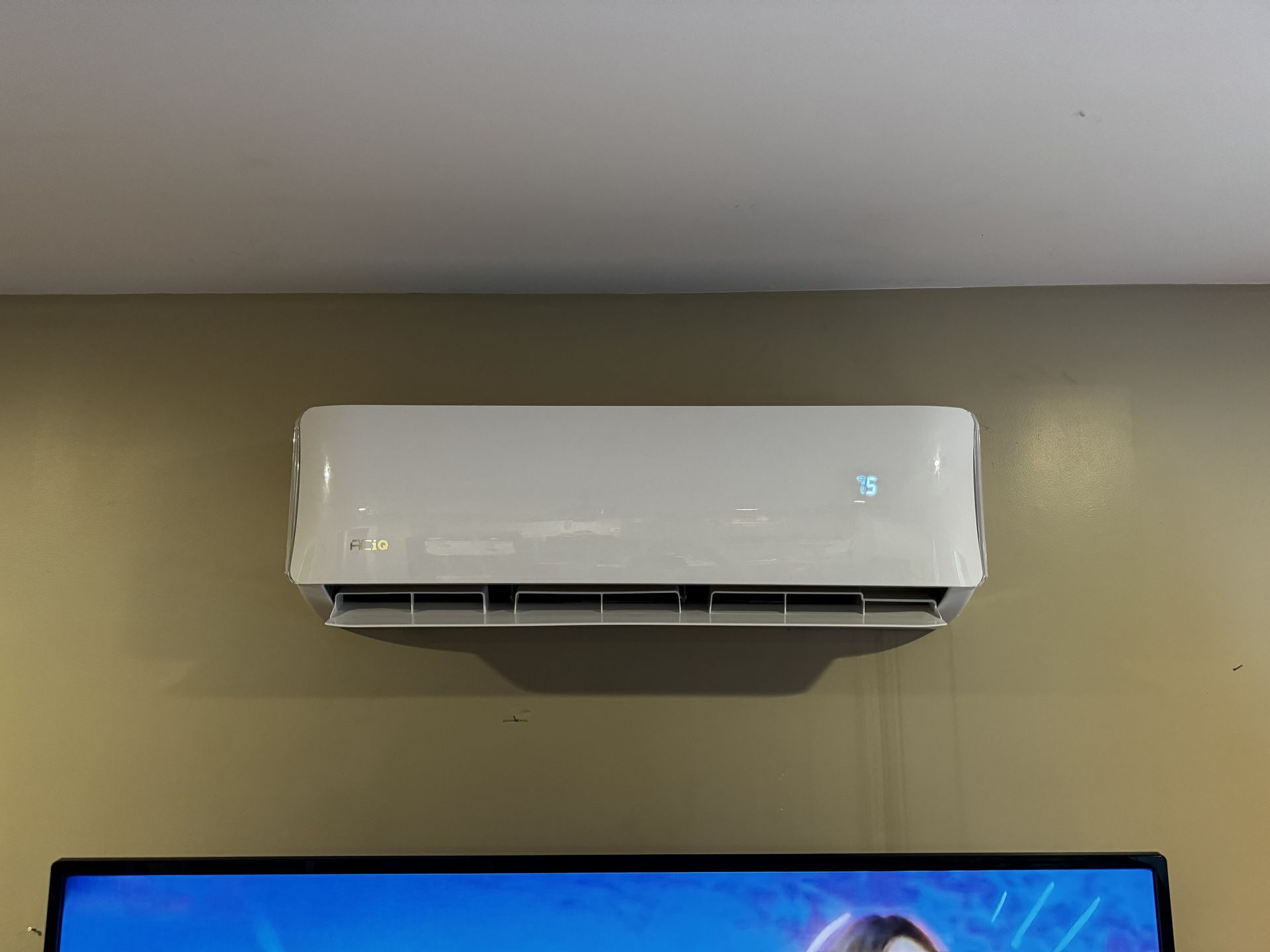 A white, rectangular air conditioning unit mounted on a beige wall above a television.