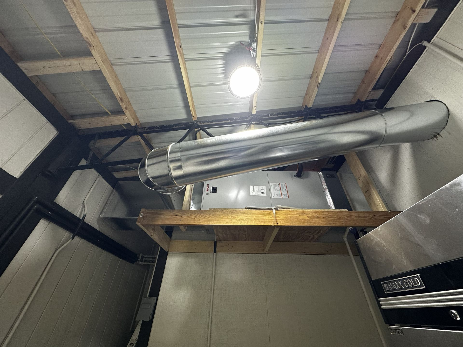 An HVAC unit mounted on a wooden frame with a metal duct extending across the ceiling under a light fixture.