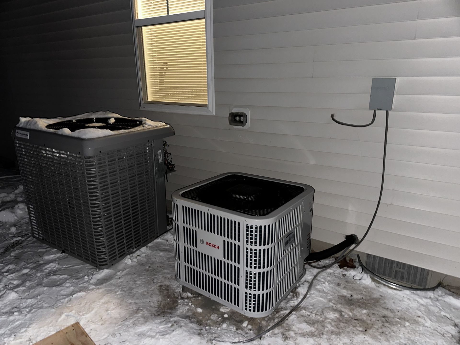Two HVAC units sit on snowy ground outside a house exterior with gray siding and an electrical disconnect box.