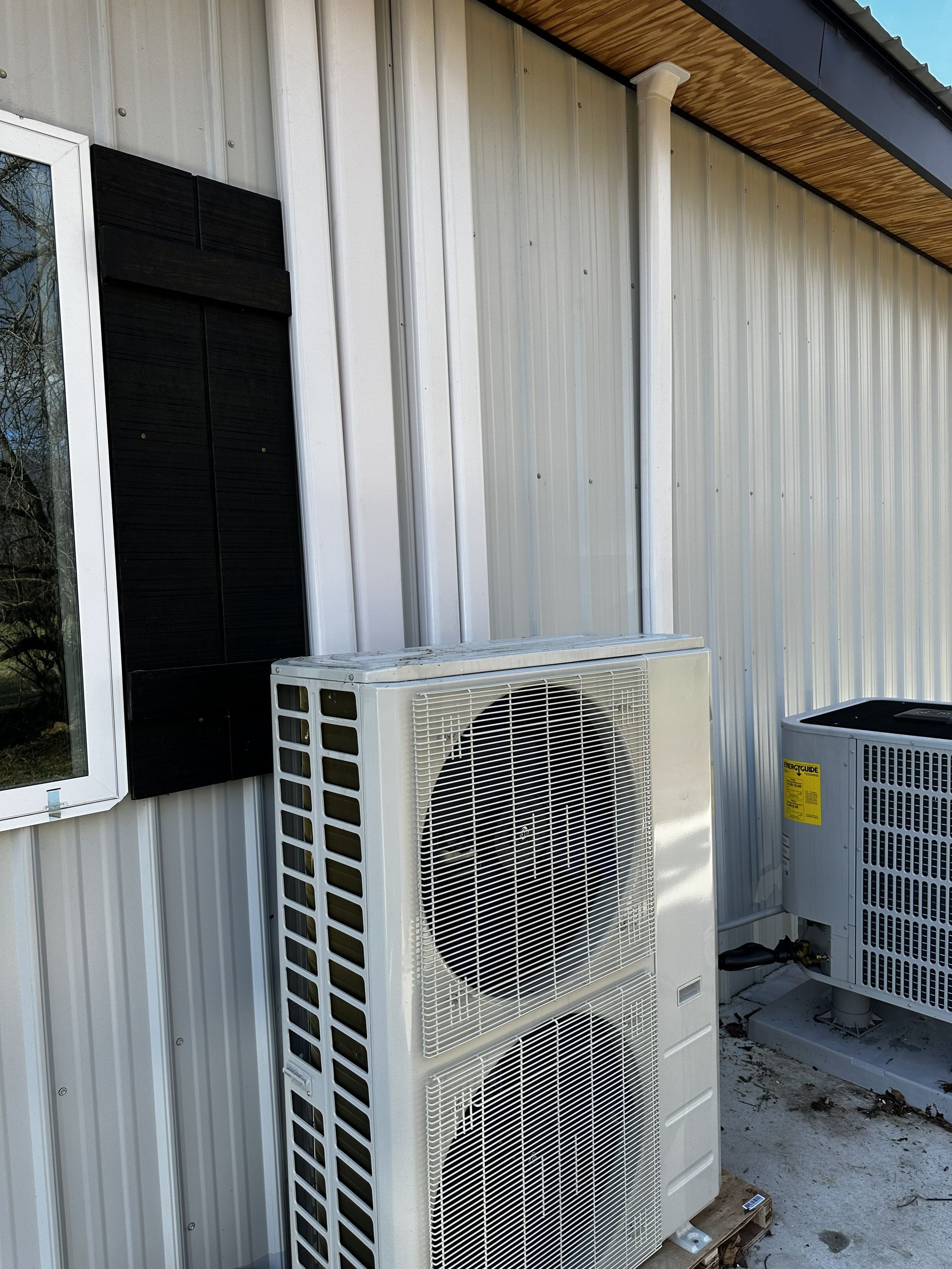 Two HVAC condenser units sit on a concrete pad against the light-colored metal siding of a building exterior.