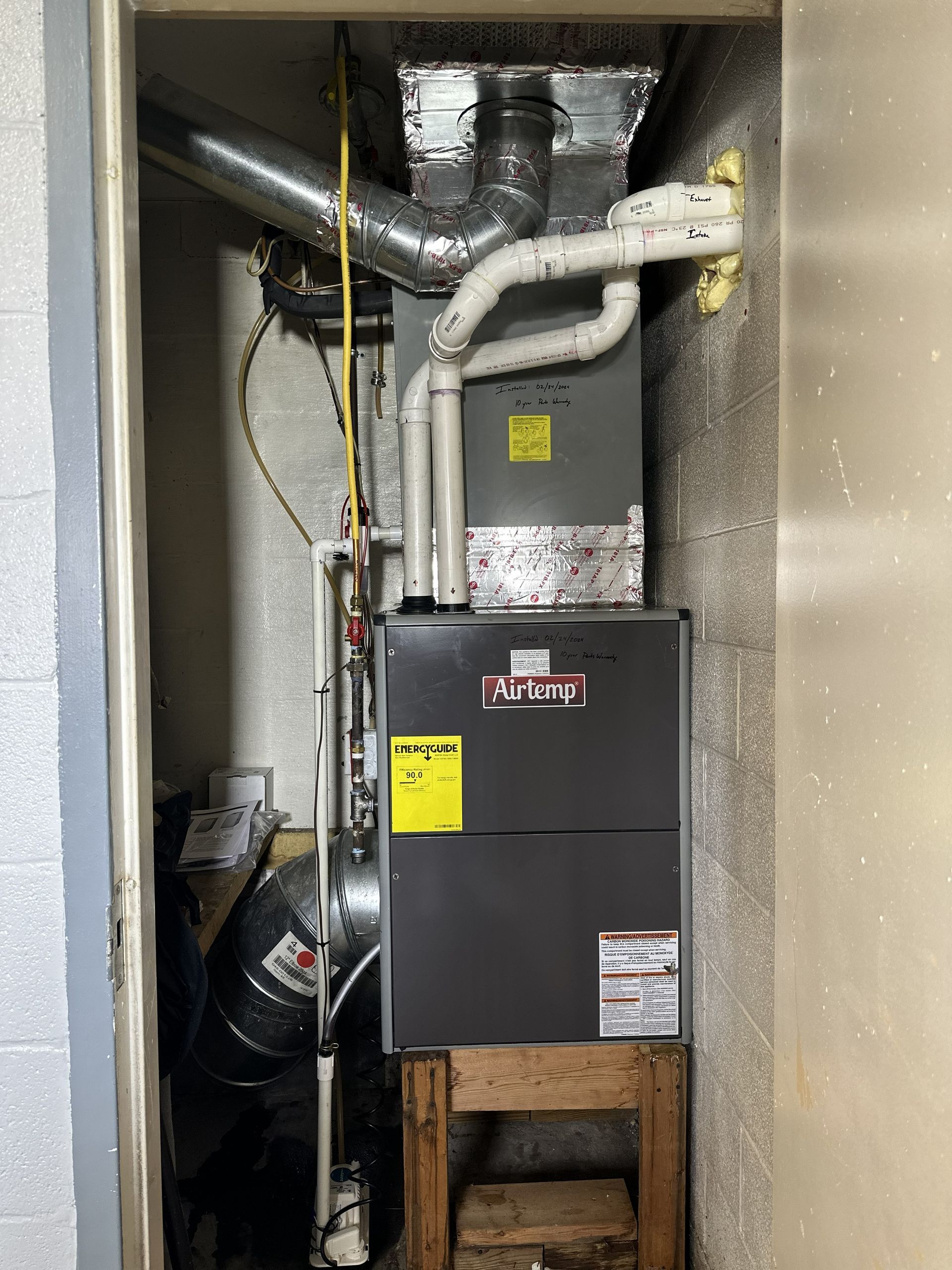 A gray vertical furnace unit housed in a small utility closet, with white PVC piping and silver ductwork connected to it.