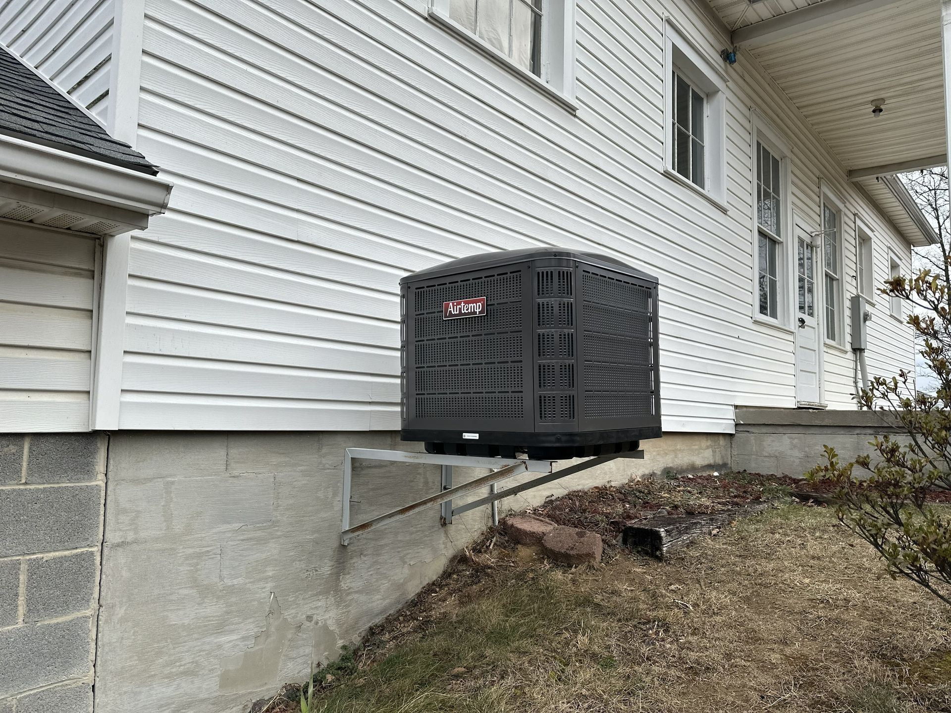A gray HVAC unit mounted on a metal bracket against the side of a white-sided house with a concrete foundation.