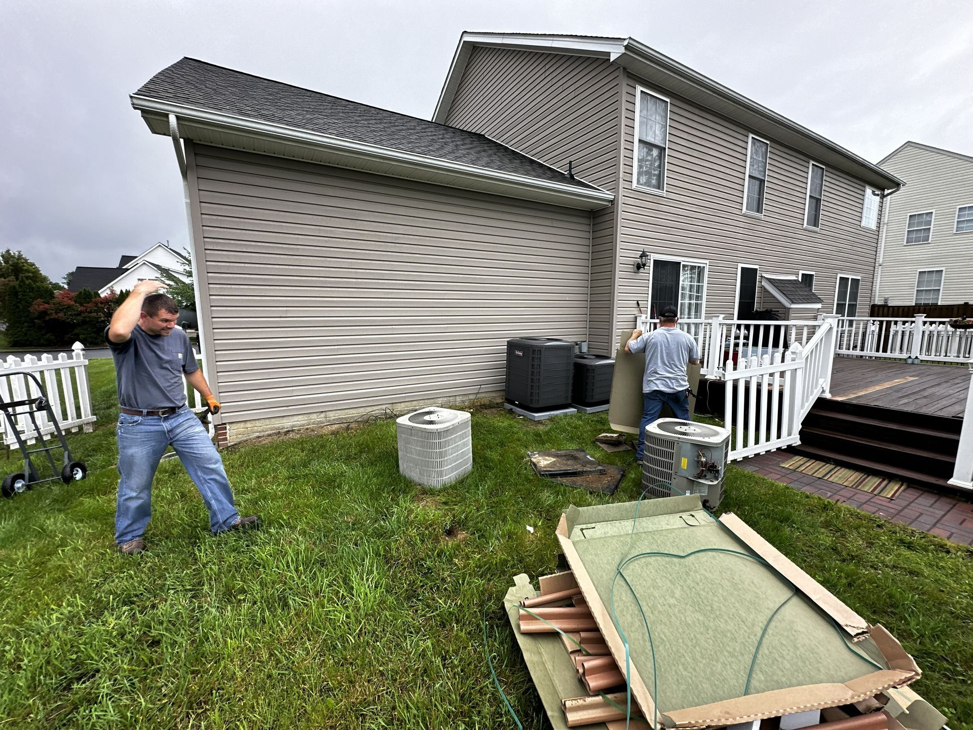 Two people move air conditioning equipment in the grass outside a house with tan siding and a white deck.