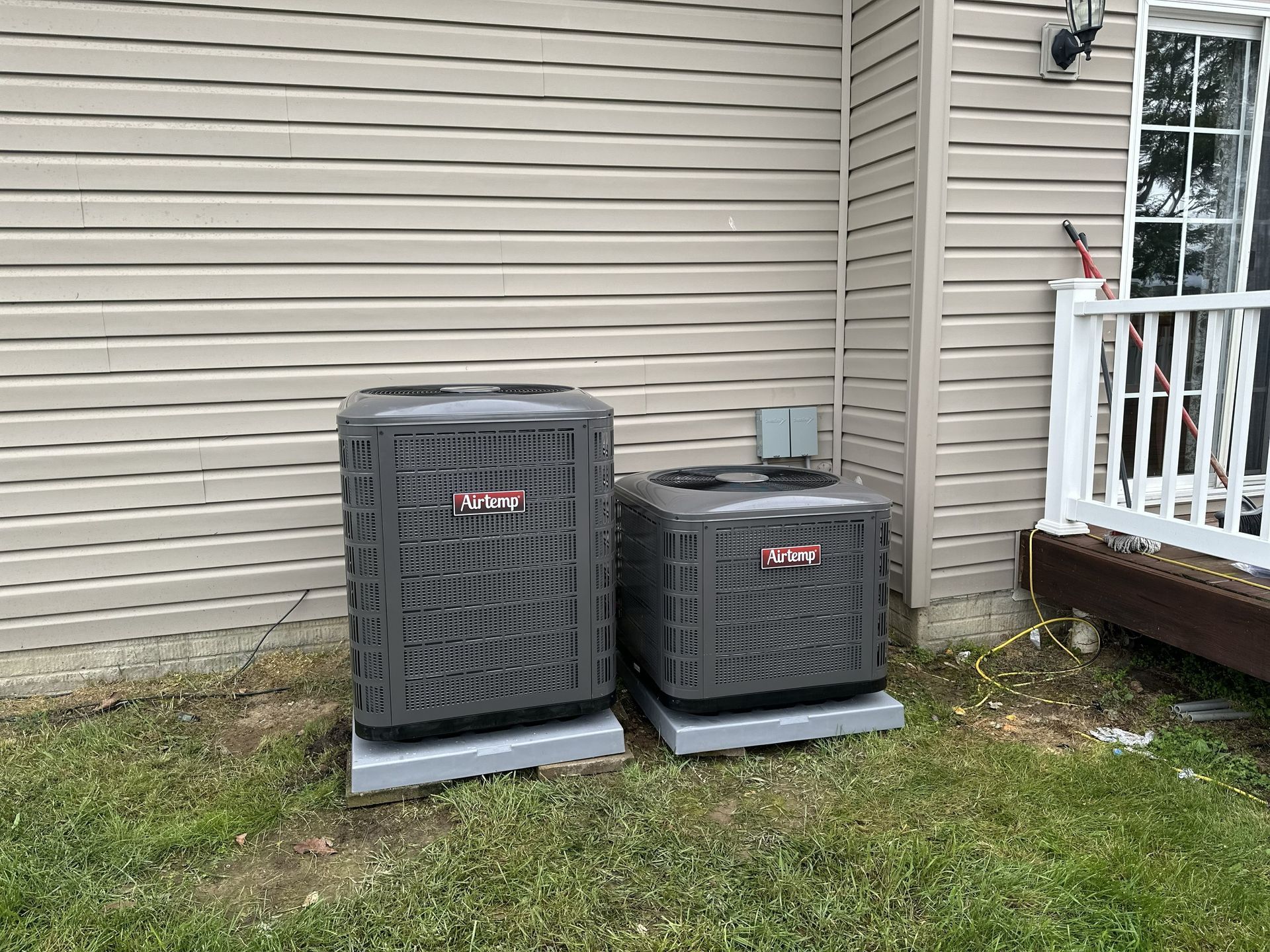 Two gray HVAC condenser units sit on concrete pads beside a beige-sided house next to a wooden porch railing.