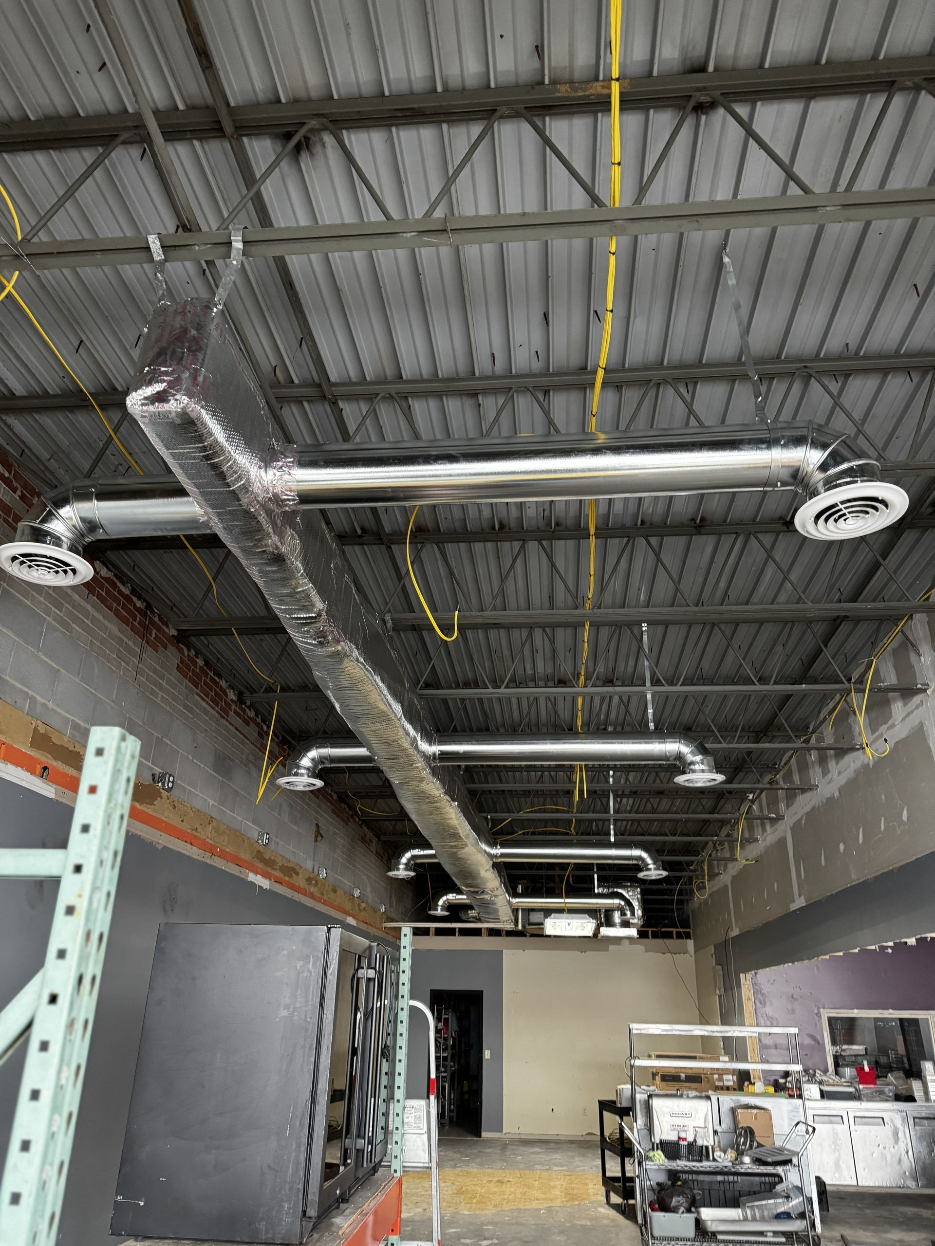 An interior view of a room under construction, featuring exposed ceiling rafters, ductwork, and hanging vents.