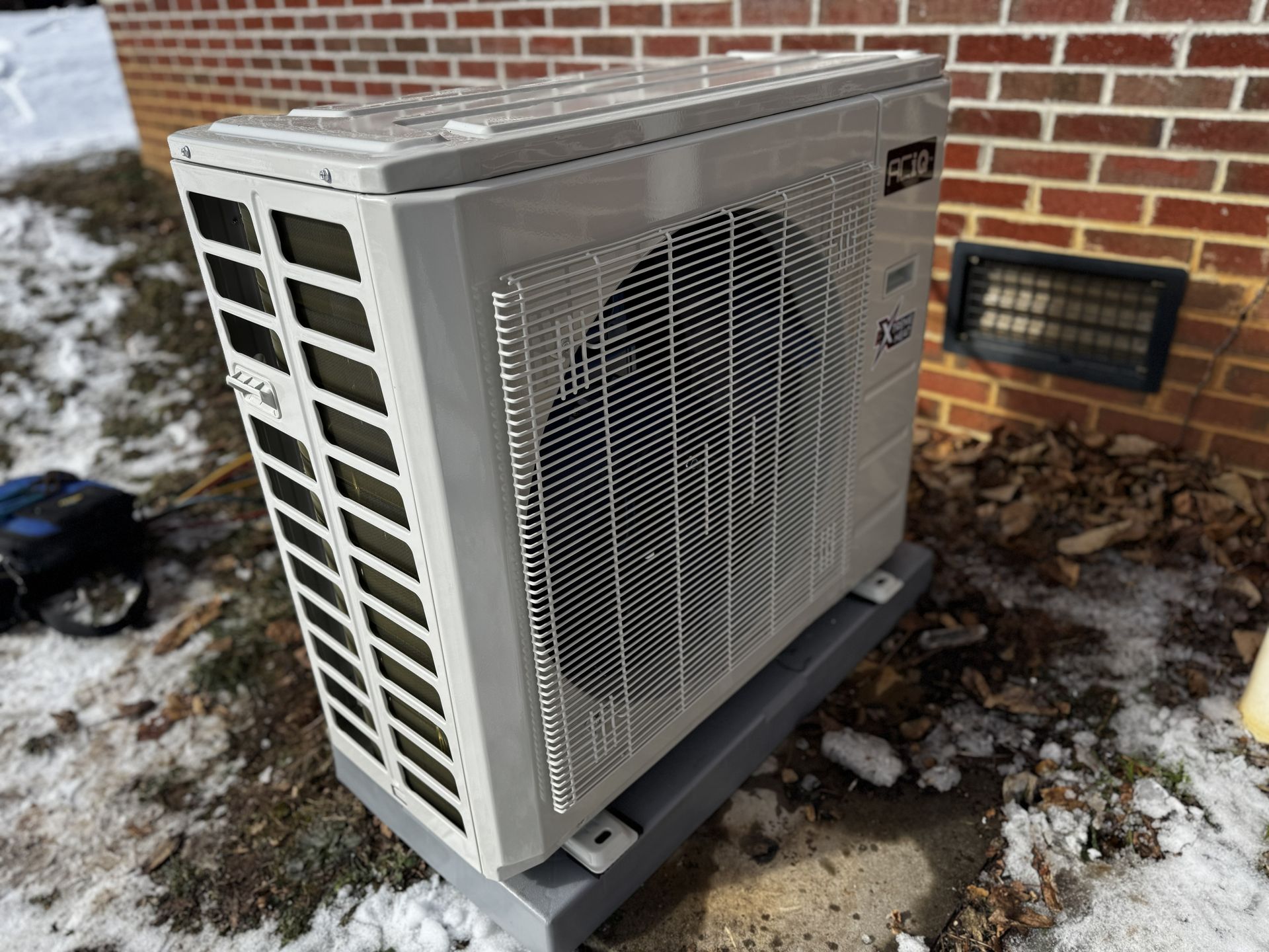 A beige outdoor heat pump unit sits on a grey plastic base against a brick wall with snow on the ground nearby.