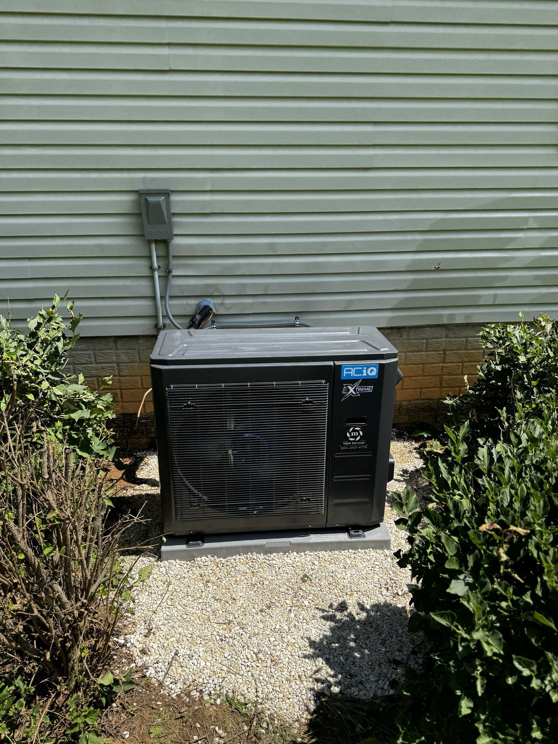 A black heat pump unit sits on a gravel patch against a wall with light green horizontal siding, flanked by green bushes.