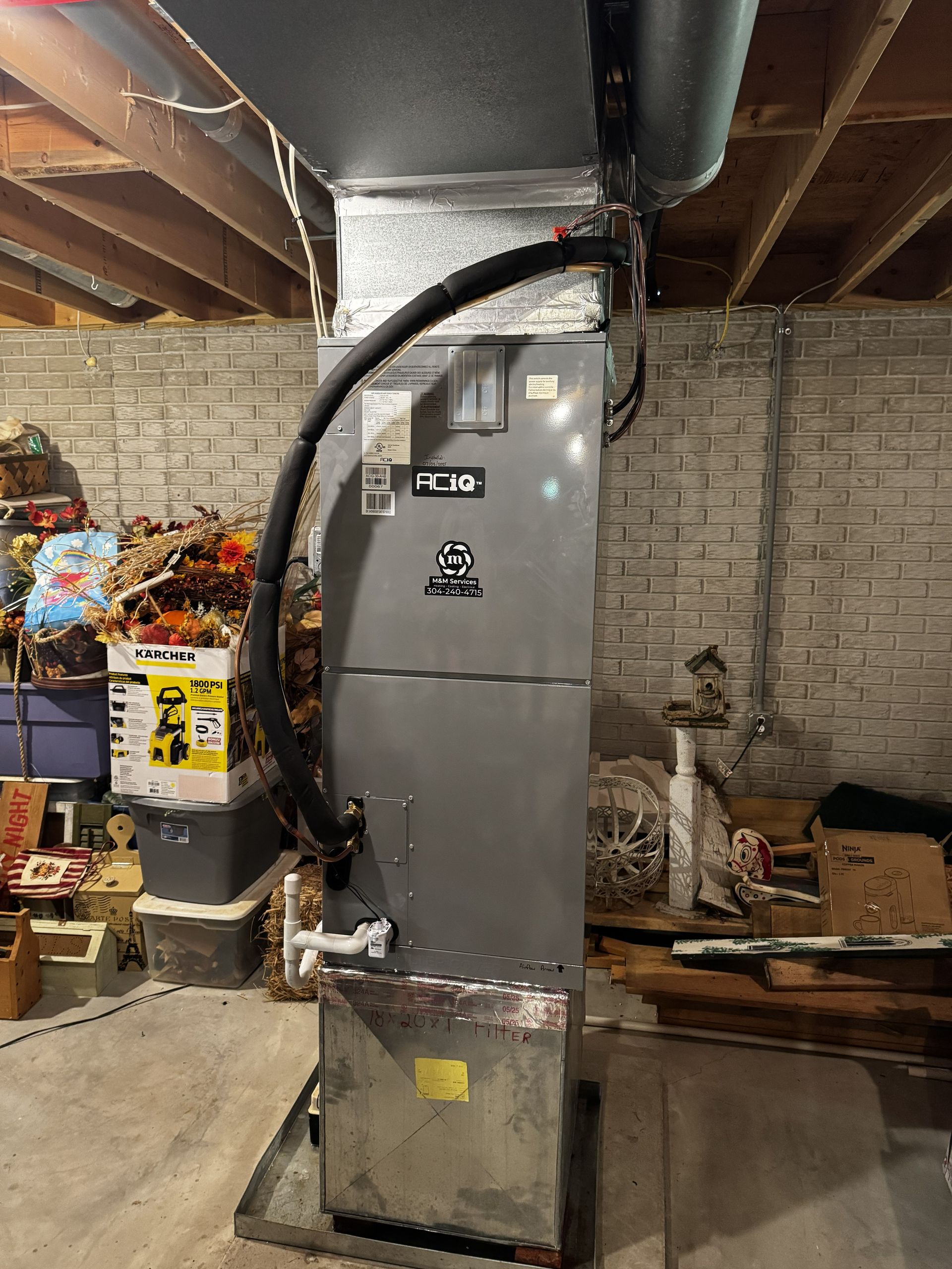 A grey HVAC air handler unit standing vertically in a basement, connected to overhead ductwork and a drain line.