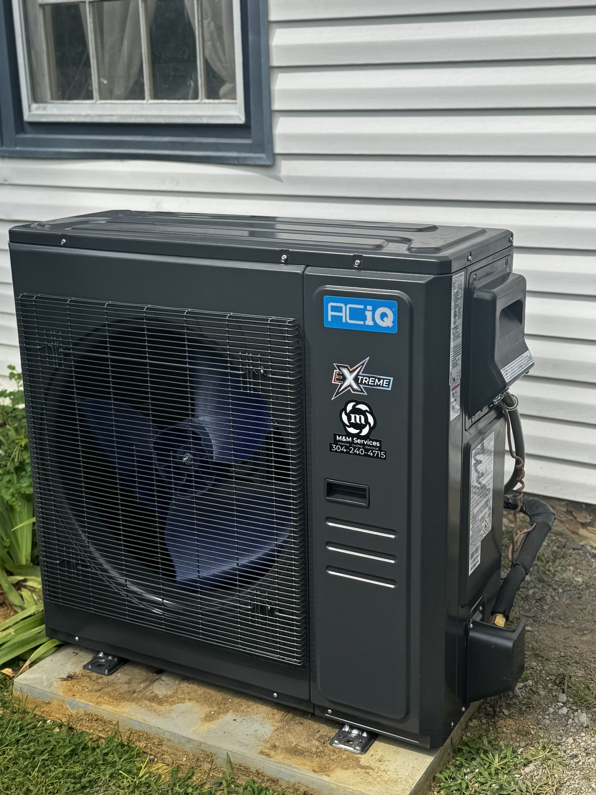A dark gray, compact outdoor HVAC condenser unit by ACIQ installed on a concrete pad against a white-sided building.