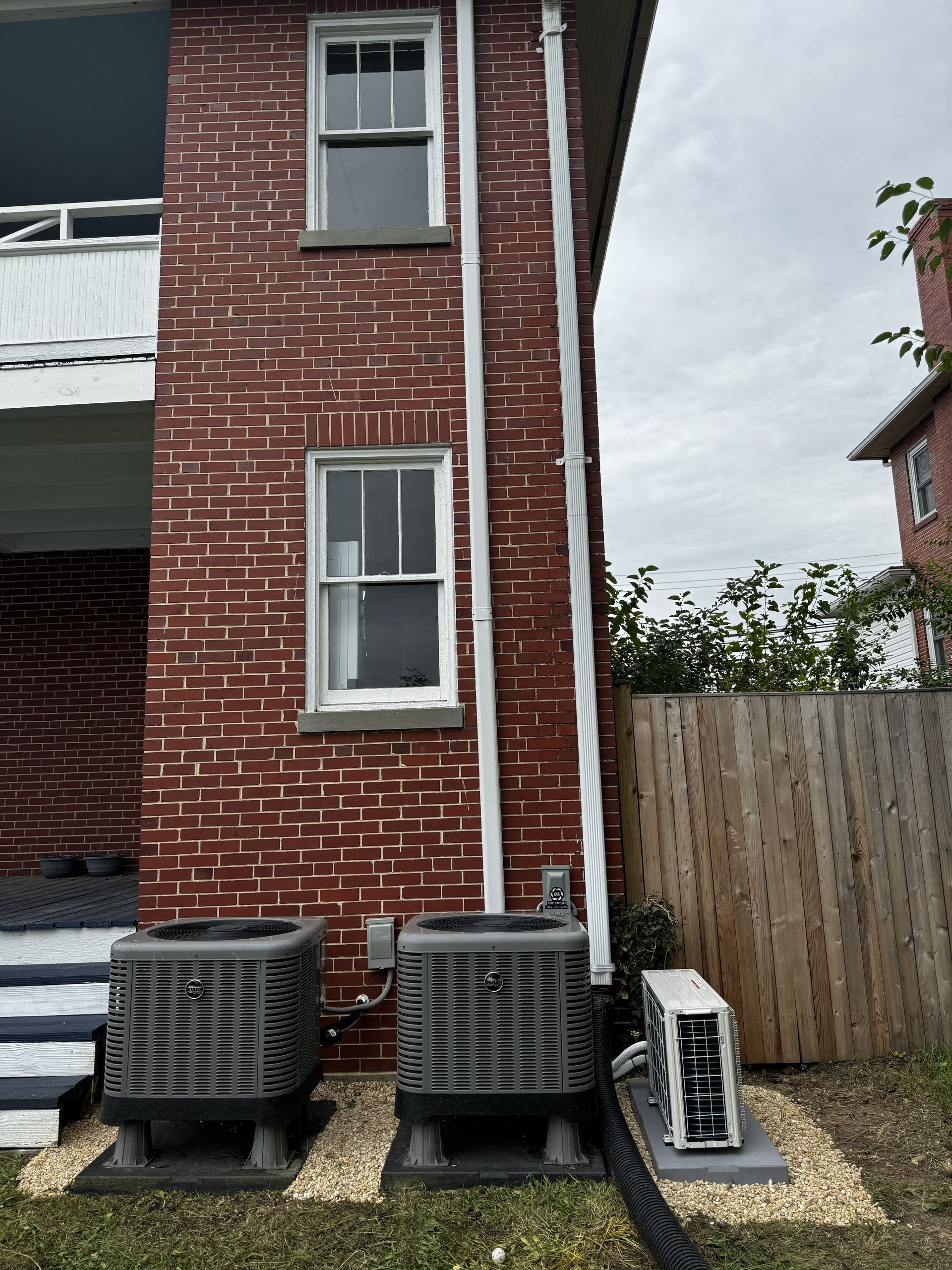 Two HVAC units and a smaller heat pump sit on gravel pads next to a brick building with white window frames and trim.