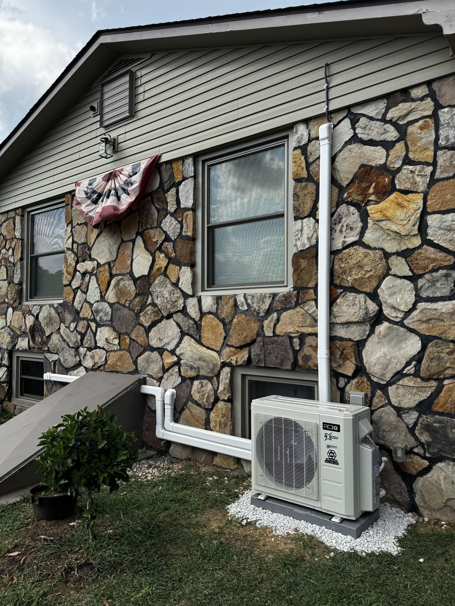 An outdoor air conditioning unit sits on a gravel base next to a stone-walled house, with white pipes running up the wall.
