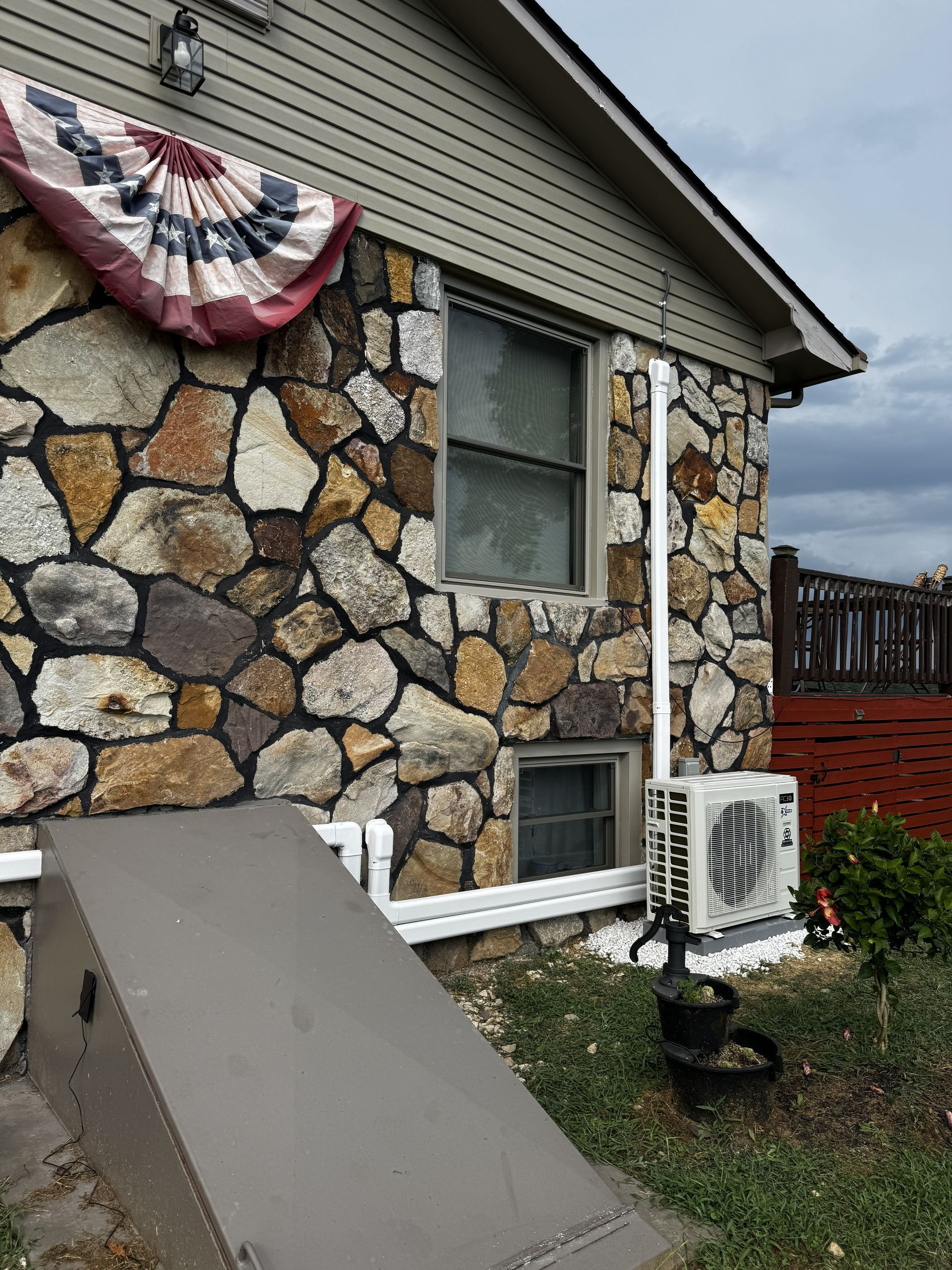 Exterior side wall of a stone house with a window, a basement bulkhead door, and an outdoor HVAC unit installed below.