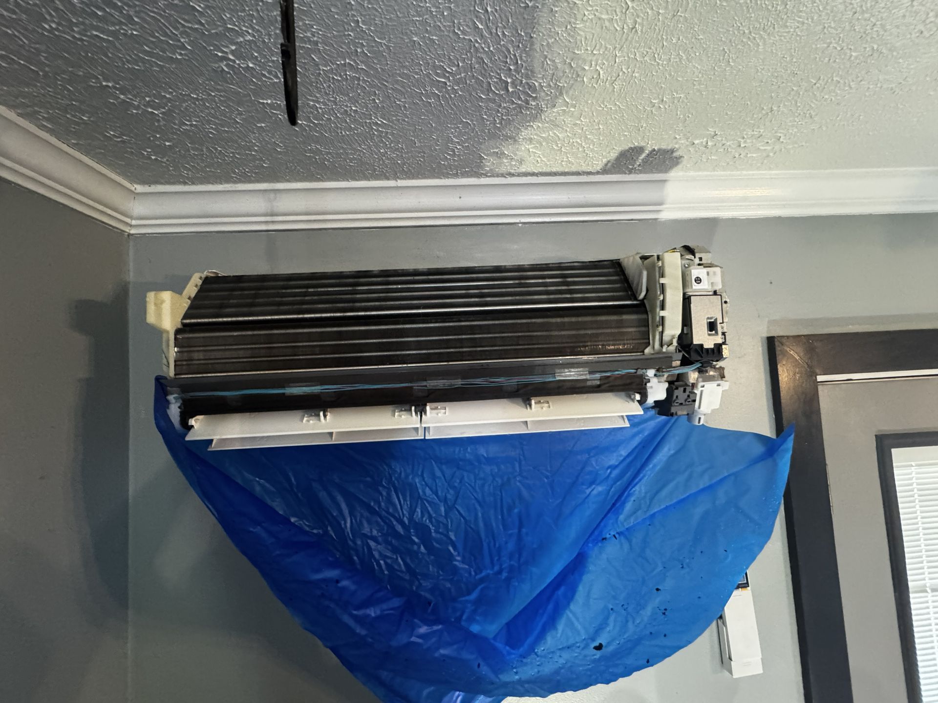 An indoor wall-mounted air conditioner unit with its front cover removed, draped with a blue plastic sheet for cleaning.