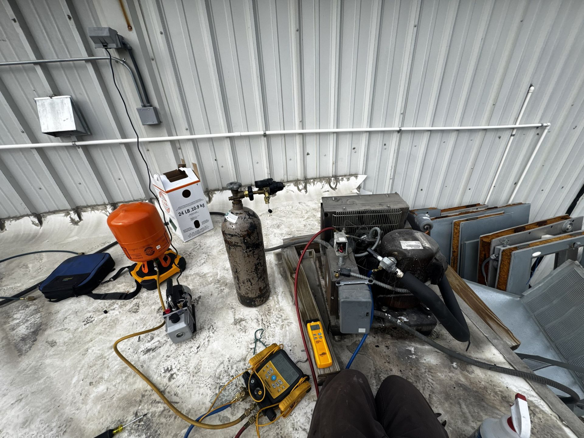 HVAC technician repairing an outdoor refrigeration unit on a roof with recovery cylinders and tools.