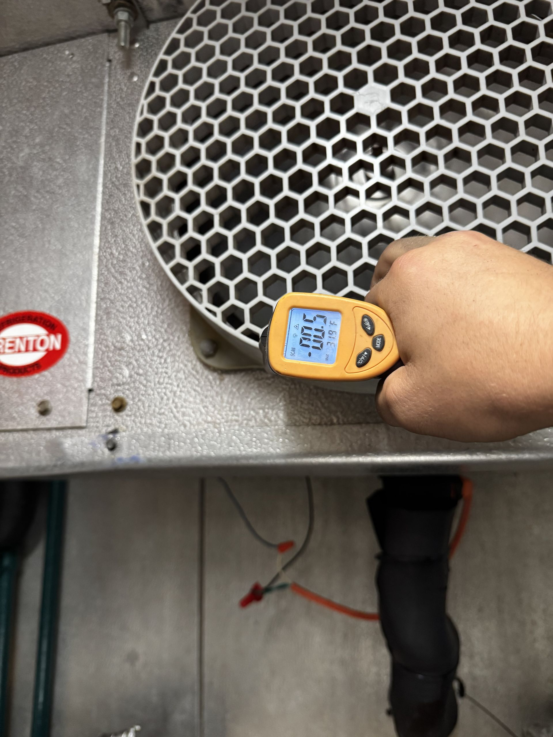 A hand holds a yellow infrared thermometer up to a large, circular metal ventilation fan, displaying a reading of 83.