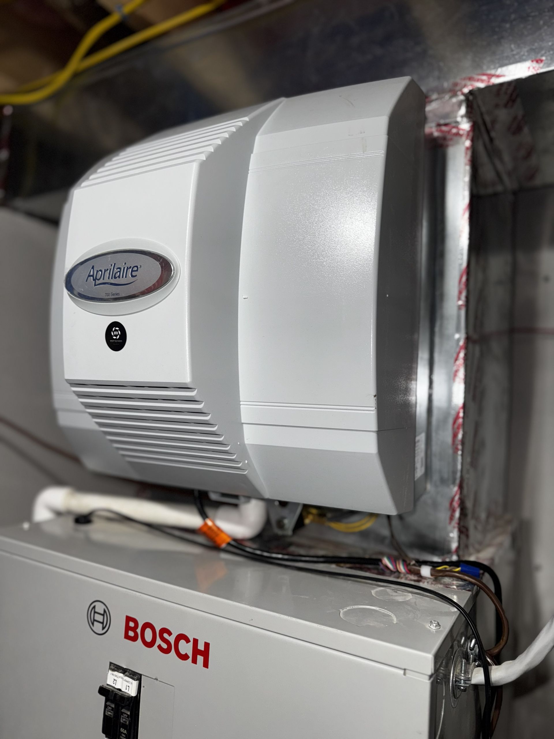 A white Aprilaire humidifier mounted on a metal duct above a Bosch furnace unit.