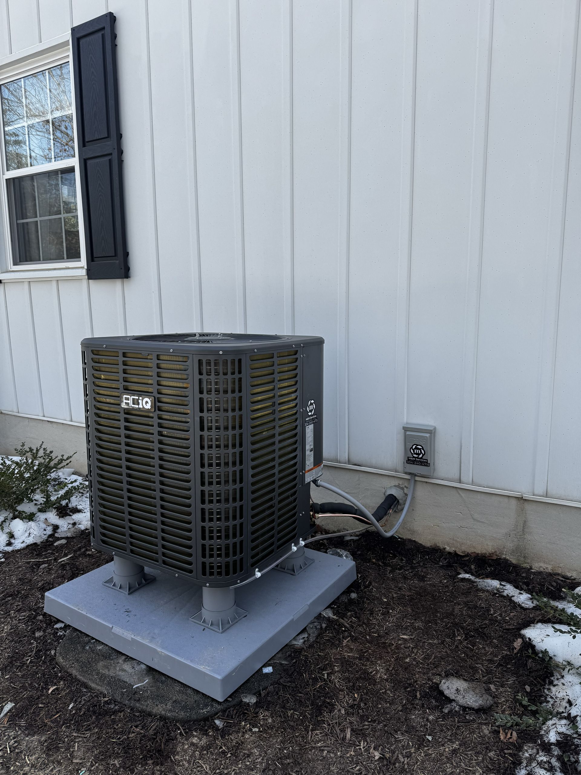 An outdoor air conditioning unit sits on a grey platform next to a white house wall, with a thin dusting of snow on ground.
