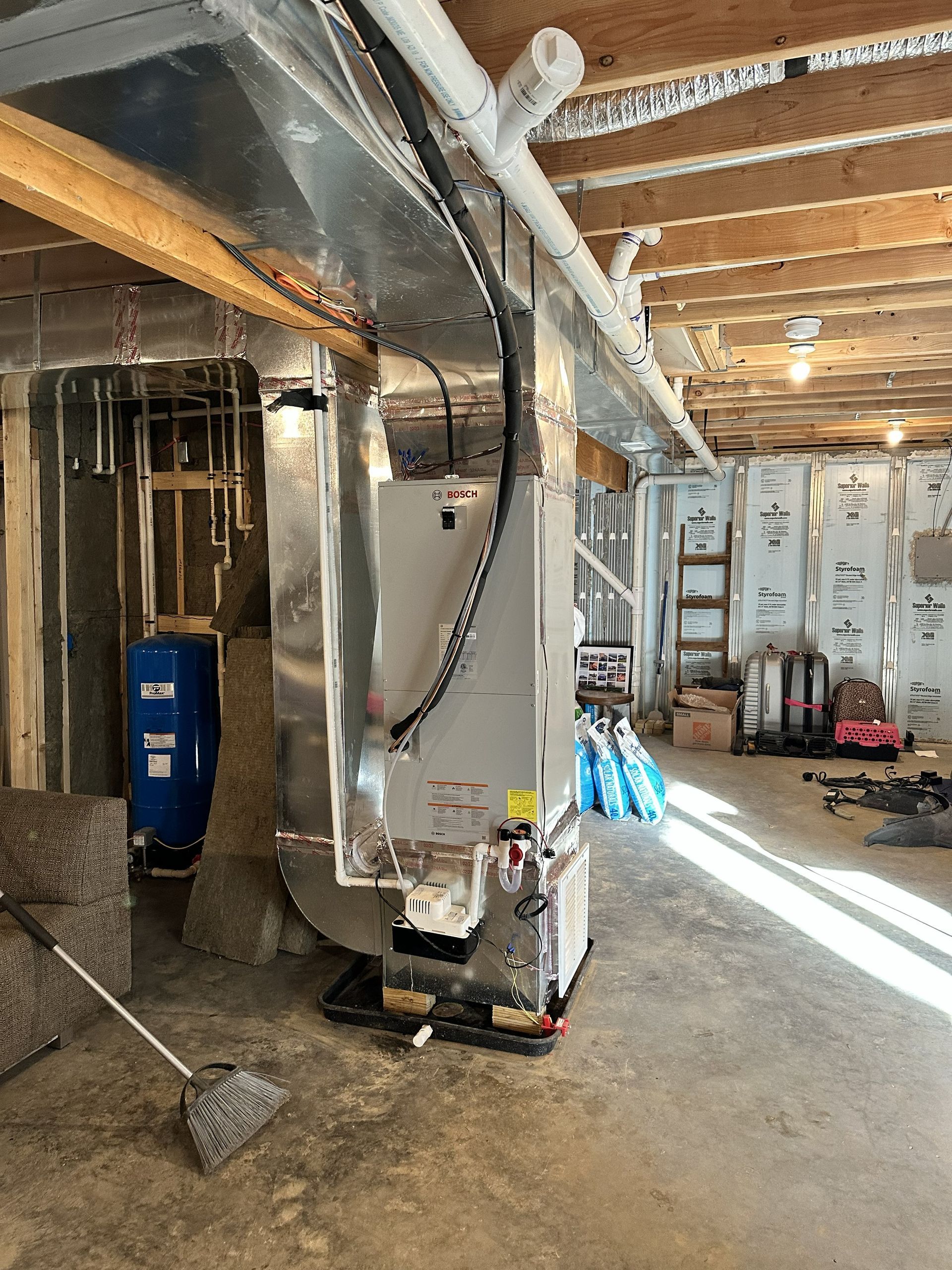 A grey HVAC unit in an unfinished basement, connected to overhead metal ductwork, next to a blue water pressure tank.